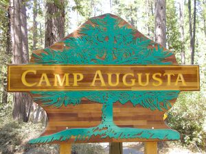 A wooden sign shaped like a tree reads Camp Augusta in yellow letters, with a green tree design carved into the wood. Pine trees and forest vegetation are visible in the background.