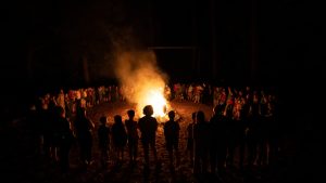 A large group of people stand in a circle around a bright campfire at night, their silhouettes illuminated by the flames—an experience made possible by every Camp Augusta donor, surrounded by darkness and towering trees.