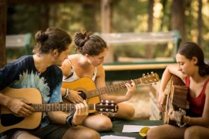Three young people sit outdoors playing acoustic guitars together, focused on their music. Trees and blurred greenery are visible in the background, creating a relaxed, natural setting.