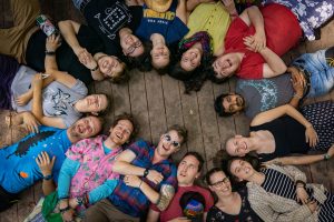 A group of people at kids summer camp lie on their backs in a circle on a wooden floor, heads together, smiling and laughing as they look up at the camera.