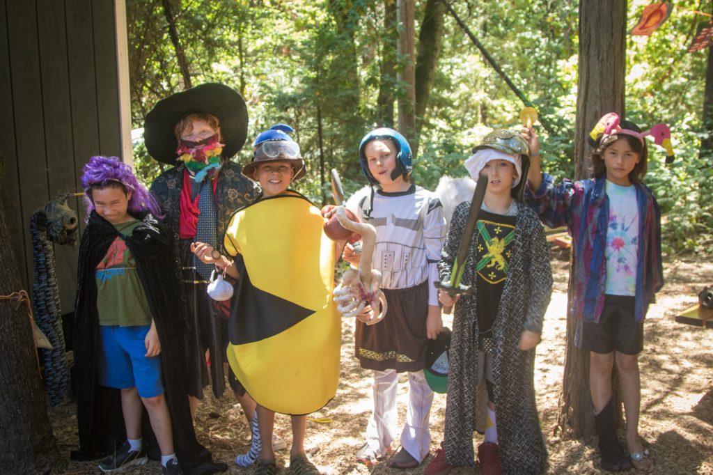 Seven kids at summer camp, dressed in colorful, creative costumes, stand together outdoors in a wooded area, smiling and posing for the camera with various props like hats, capes, and shields.