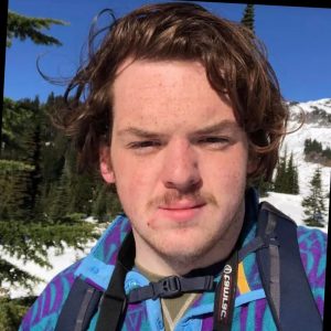 A young person with wavy brown hair stands outdoors in a snowy, mountainous area, wearing a blue patterned jacket and a backpack with a camera strap over their shoulder. Evergreen trees and snow are visible in the background.