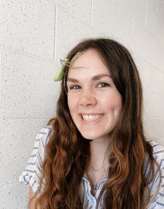 A smiling young woman with long brown hair and a striped shirt has a green praying mantis sitting on her forehead, standing against a light-colored brick wall.