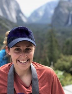 A smiling person wearing a navy blue cap and a red shirt stands outdoors with a blurred background of mountains, trees, and a waterfall.