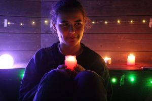A person sits in dim lighting, holding a glowing candle with both hands. Colorful candles and string lights illuminate the dark wooden background, creating a warm and cozy atmosphere.