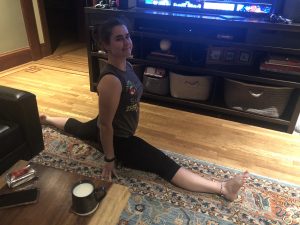 A person in a dark tank top and leggings is doing a split on a wooden floor in a living room, smiling at the camera. A TV, shelves, and a coffee table with a mug of milk are visible in the background.