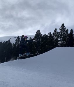 A skier wearing a helmet and goggles is airborne after jumping off a snow-covered slope, with dark evergreen trees and a cloudy sky in the background.