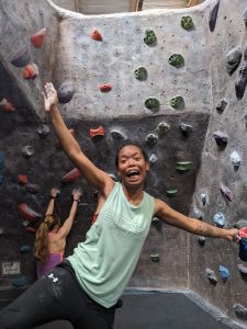 A smiling person in a green tank top enthusiastically poses in front of a rock climbing wall, while another person climbs in the background.