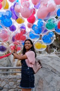 A smiling woman in a pink shirt and denim overalls holds a large bunch of colorful balloons outdoors, standing in front of rocks and trees.