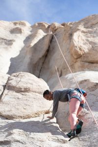 A person in climbing gear ascends a steep, rocky cliff outdoors, secured by a rope anchored above them under a clear blue sky.