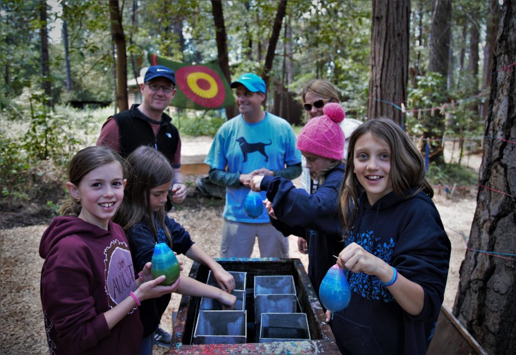 A group of children and adults outdoors in a wooded area, smiling and making crafts with colored balloons and water at a table. Trees and a colorful target are in the background.