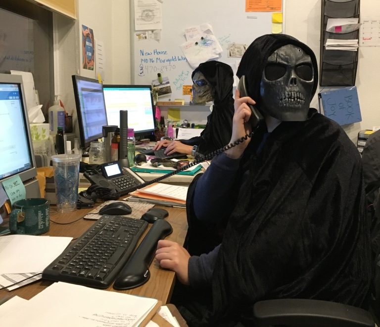 Two people wearing black cloaks and skull masks work at desks with computers and paperwork in an office; one is on the phone, and the other types on a keyboard. A whiteboard and office supplies are in the background.