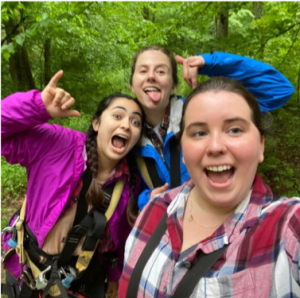 Three young women smile and make playful faces while posing for a selfie in a lush, green forest. They are wearing plaid shirts and harnesses, suggesting they may be on an outdoor adventure.