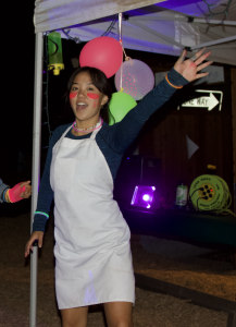A person wearing a white apron, blue long-sleeve shirt, and neon face paint raises one arm and smiles at an outdoor nighttime event decorated with glow sticks and colorful balloons.