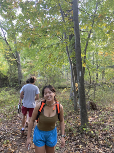Two people are hiking on a leafy trail in a green forest. The person in front, smiling, wears a brown tank top, blue shorts, and an orange backpack. The person behind wears a gray shirt and holds their shoes.