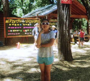 A young person in sunglasses stands with arms crossed and making a peace sign in front of an “Activity Sign-Ups” board at an outdoor camp, surrounded by trees and other people in the background.