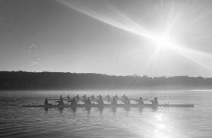 A black and white photo of a rowing team gliding across a calm lake at sunrise, with sunlight reflecting on the water and a misty treeline in the background.