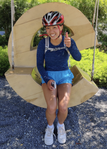 A smiling person wearing a red bike helmet, blue shirt, and blue shorts sits on a large wooden swing shaped like a letter S, giving a thumbs-up gesture outdoors.
