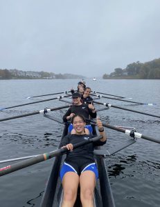 A rowing team of eight people sits in a boat on a calm river, all smiling and giving thumbs up. The team is dressed in matching athletic gear, with trees and buildings visible on the misty shoreline in the background.