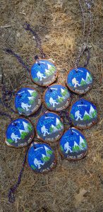 Nine painted wooden medallions with braided strings lay on pine needles. Each has a blue background, white figure, trees, and mountain, and a different name with “Circle 2019” written at the bottom.