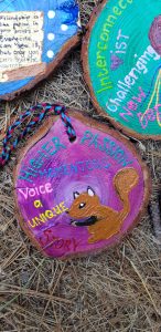 A painted wood slice features a brown squirrel, colorful words like HIGHER PASSION, MOMENTOUS, Voice, UNIQUE, and STORY, with a braided string attached, resting on straw among other decorated wood slices.
