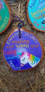 A painted wooden slice with a colorful unicorn and the words Empathic Villager catching the WOAH and Failing towards Meaning, set on straw. A braided cord is attached to the top.