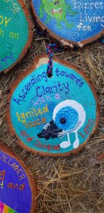 A painted wood slice with an illustrated blue eye holding a black megaphone. The text reads: Ascending towards Clarity with a Ignited Voice and Unique Lens. The wood is resting on straw with other decorated slices nearby.