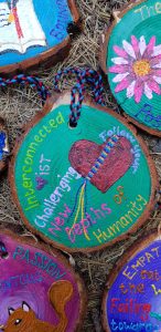 A painted wooden disc shows a heart tied with colored strings and words: Interconnected, Mist, Challenging, New Depths of Humanity, and Following. Surrounding discs have colorful art and text.