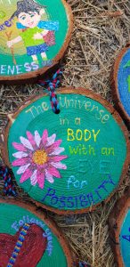 A painted wooden slice features a large pink flower and colorful text reading, The Universe in a BODY with an EYE for POSSIBILITY, lying on straw among other decorated wood slices.