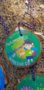 A painted wood slice shows a smiling child with short hair, a backpack, and a yellow stick. The text reads: Yellow Heart Metamorphic Map Adventure of Wholeness. The decoration hangs on a colorful braided cord.