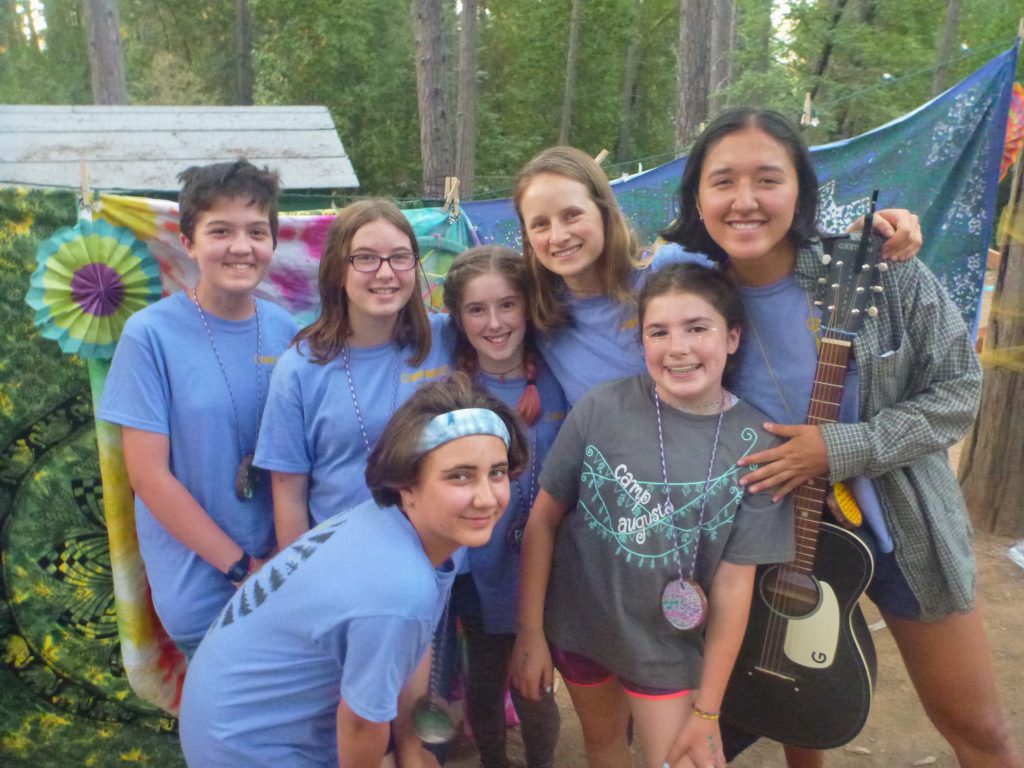 A group of six kids and one adult, all smiling and wearing blue shirts, pose outdoors in front of colorful fabric. One person holds a black acoustic guitar. Tall trees and a wooden structure are in the background.
