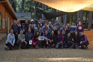 A group of women pose and smile together outdoors on wood chips, in front of a wooden building surrounded by trees, under tarps providing shade.
