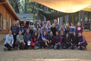 A group of about 30 women pose and smile together outdoors under shade cloths, with trees and a wooden cabin in the background. Most are dressed casually in jackets and athletic wear.