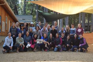 A group of around 35 women pose and smile outdoors under shade cloths, in front of a wooden building surrounded by tall trees. Some kneel or sit in front, while others stand behind.