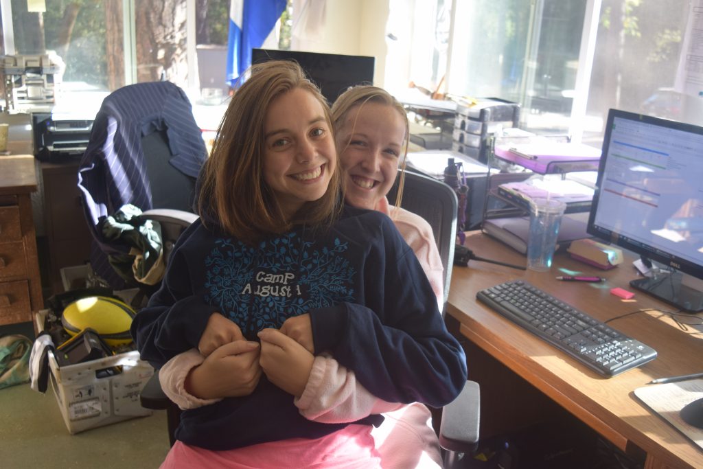 Two young women are smiling and sitting together in an office chair, one sitting on the others lap. Sunlight streams in from large windows in the background, and a computer and office supplies are on the desk beside them.