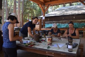 Four women work on arts and crafts at a wooden table in a covered outdoor area surrounded by trees and cabins. They appear to be focused and smiling, enjoying the activity together in a rustic setting.