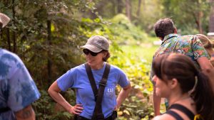 A woman wearing sunglasses, a gray cap, and a blue shirt stands outdoors with hands on hips, smiling and talking to others in a sunlit, wooded area. Several people are nearby, partially visible.