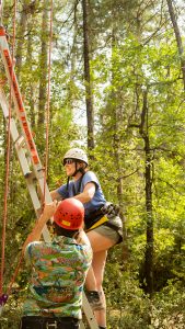 A woman wearing a helmet and harness climbs a ladder in a forested area while a person in a red helmet and colorful shirt steadies the ladder for her. Sunlight filters through the trees in the background.