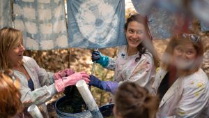 Four women wearing paint-splattered lab coats work together on a tie-dye project outdoors, smiling and chatting beneath hanging blue and white patterned fabric.