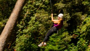 A person wearing a helmet and harness glides on a zip line through a forested area, surrounded by green trees.