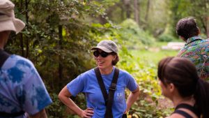 A woman wearing sunglasses, a hat, and a blue shirt stands outdoors in a forested area, smiling and talking with a group of people. The background is lush and green.