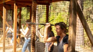 A woman stands behind a net, holding a spear above her shoulder as if preparing to throw it. Other people in the background also appear engaged in similar activities at an outdoor range. Sunlight illuminates the scene.
