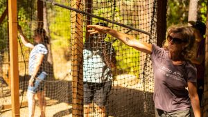 A woman wearing sunglasses reaches toward a netted structure outdoors, while two other people stand nearby in a wooded area with sunlight filtering through the trees.