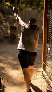 A person in a gray shirt, black shorts, and a cap throws an axe toward a wooden target at an outdoor axe-throwing range on a sunny day.