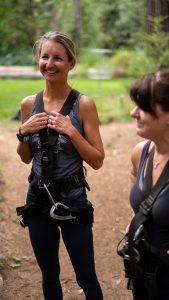 Two women wearing harnesses and athletic gear stand outdoors on a dirt path, smiling and preparing for an outdoor adventure activity, surrounded by greenery and trees.