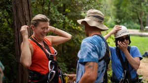 Three adults wearing harnesses and helmets prepare for an outdoor adventure activity among trees. One woman in orange smiles, a man in a hat faces her, and another person in blue adjusts their helmet while laughing.