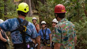 A group of people wearing helmets and harnesses stand together in a wooded area, smiling and listening to two guides in colorful shirts and helmets.