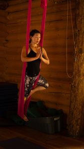 A woman practices aerial yoga indoors, balancing in a pink hammock with hands in prayer position. She stands on one foot, wearing black and white leggings and a black tank top, against a wooden wall background.