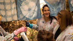 Three women wearing paint-splattered clothing and gloves smile and talk while holding up indigo-dyed fabric squares drying on a clothesline outdoors. Sunlight filters through the trees in the background.