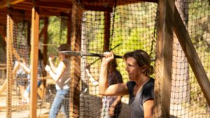 A woman holds a spear, preparing to throw it at an outdoor activity range, with other people practicing in the background, surrounded by wooden structures and netting for safety.